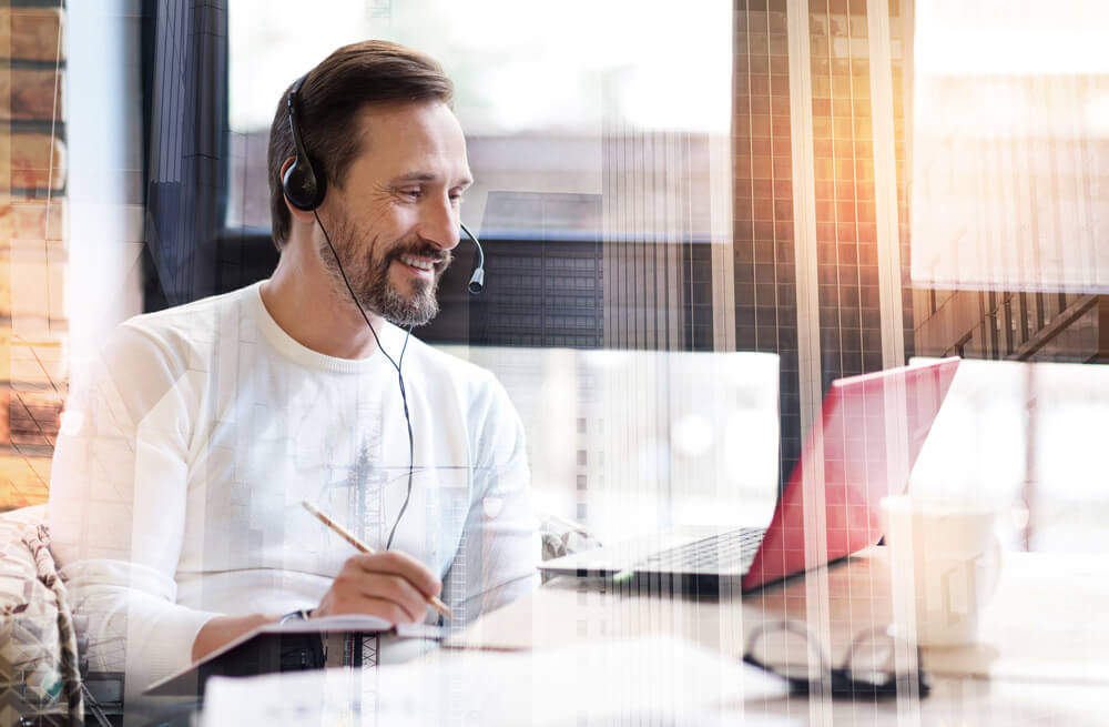 homem sorridente com fones de ouvido e microfone olhando para o laptop e escrevendo em bloco de anotações
